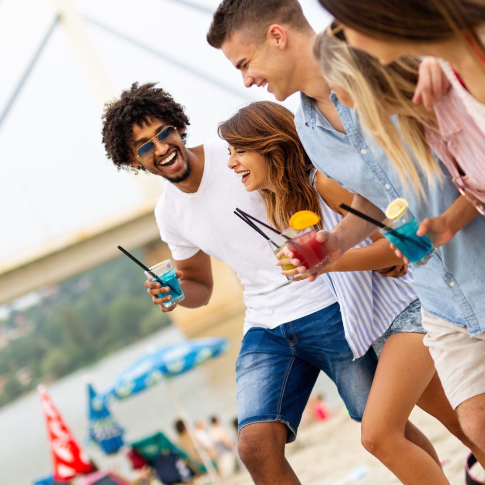 Happy friends group having fun at beach party drinking cocktail at sunset. Summer joy and friendship concept with young people on vacation